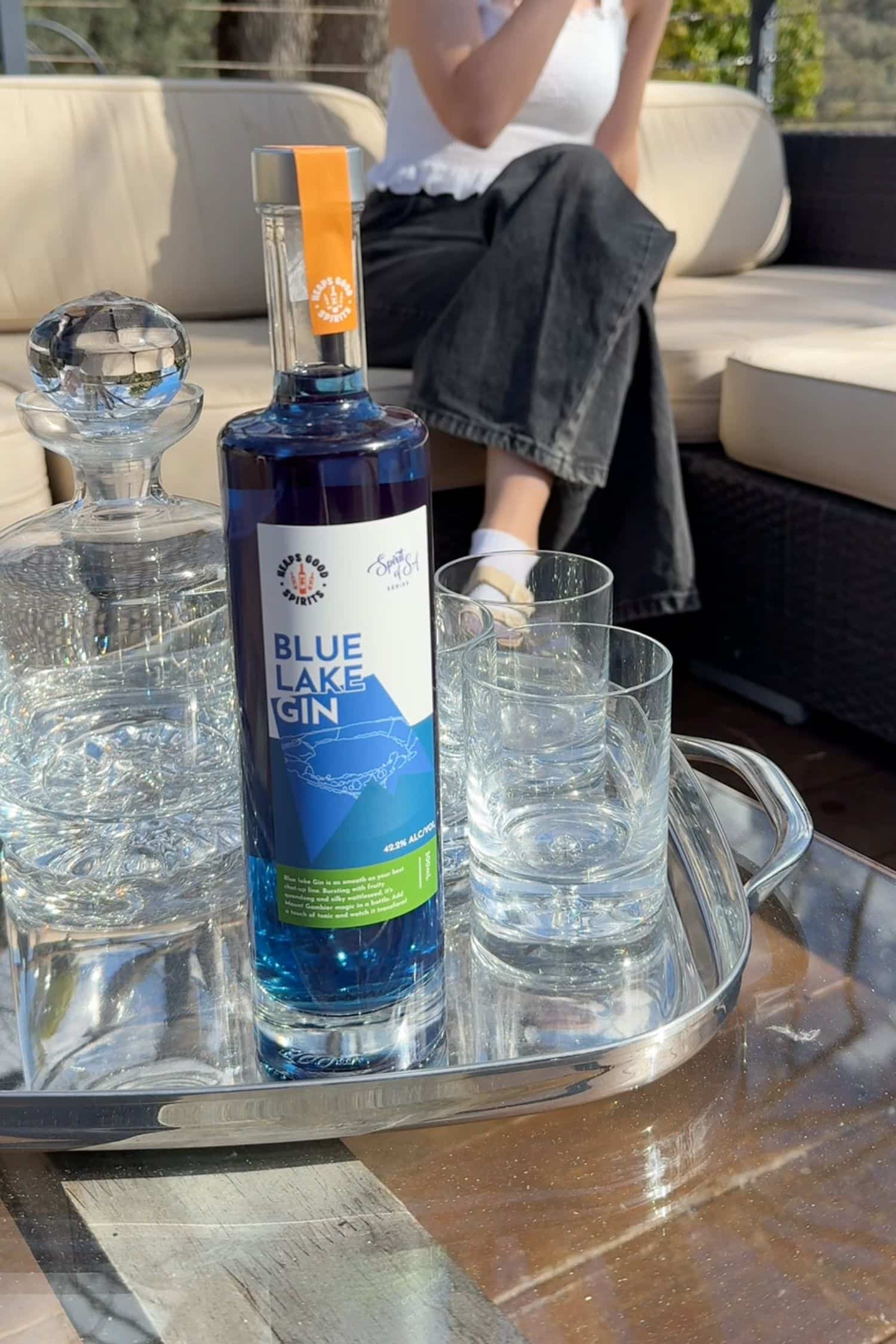 Blue Lake Gin bottle on silver tray outdoors with glasses, bright blue spirit glowing in natural sunlight.