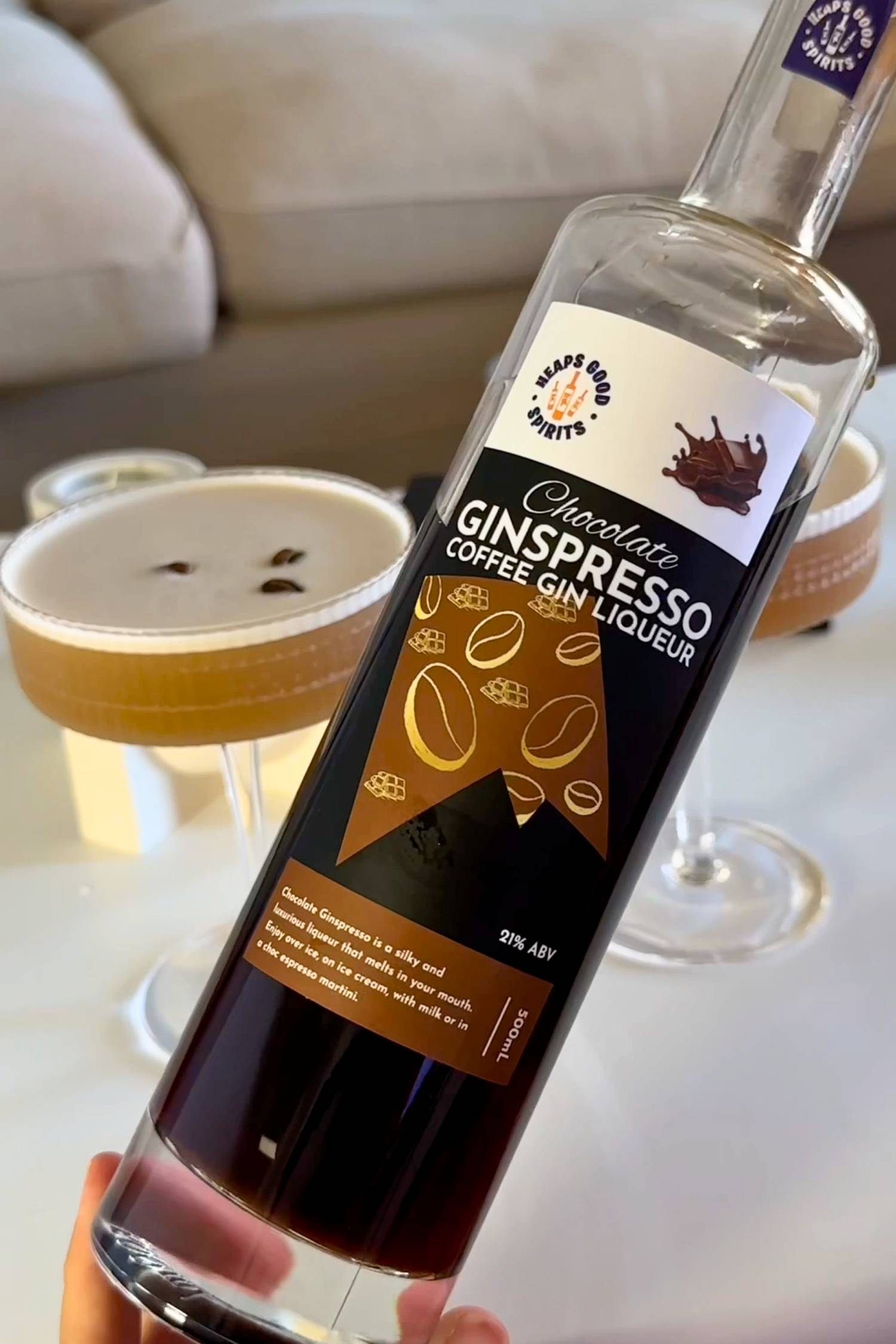 Bottle of Chocolate Ginspresso coffee gin liqueur held up with freshly made espresso martini cocktails in the background.