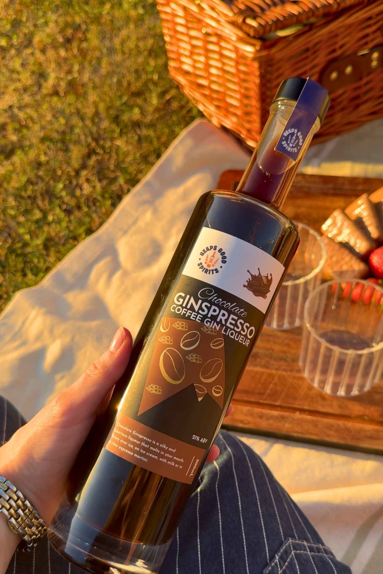 Chocolate Ginspresso coffee gin liqueur held in hand at an outdoor picnic, with TimTam biscuits and strawberries in the background.