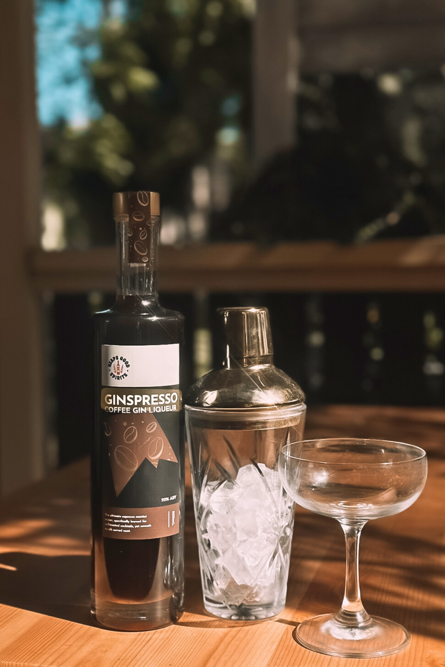 Bottle of Ginspresso coffee gin, cocktail shaker with ice, and glass on a wooden surface.