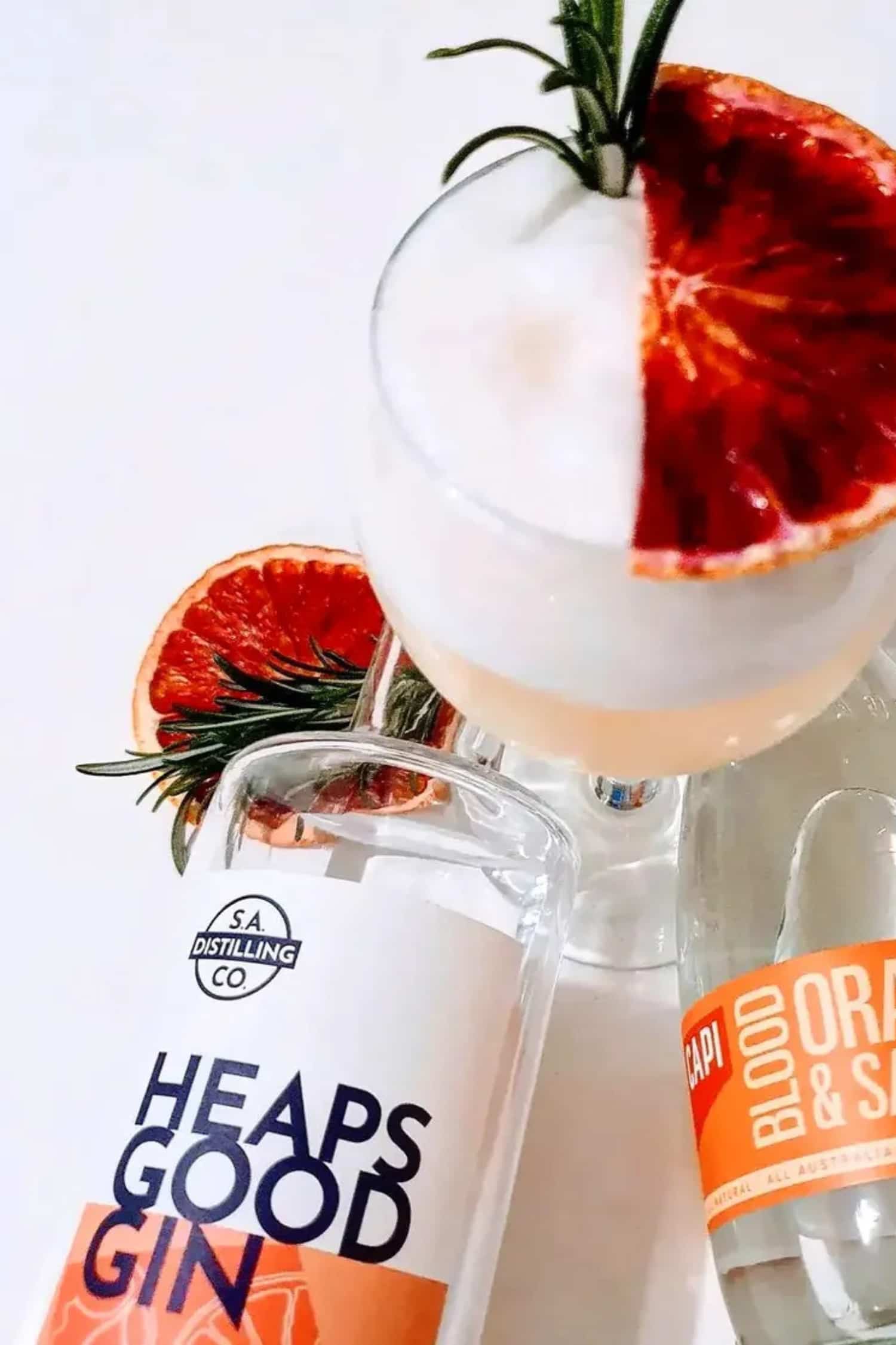 Cocktail with a slice of blood orange and rosemary, featuring a bottle of Heaps Good Gin.