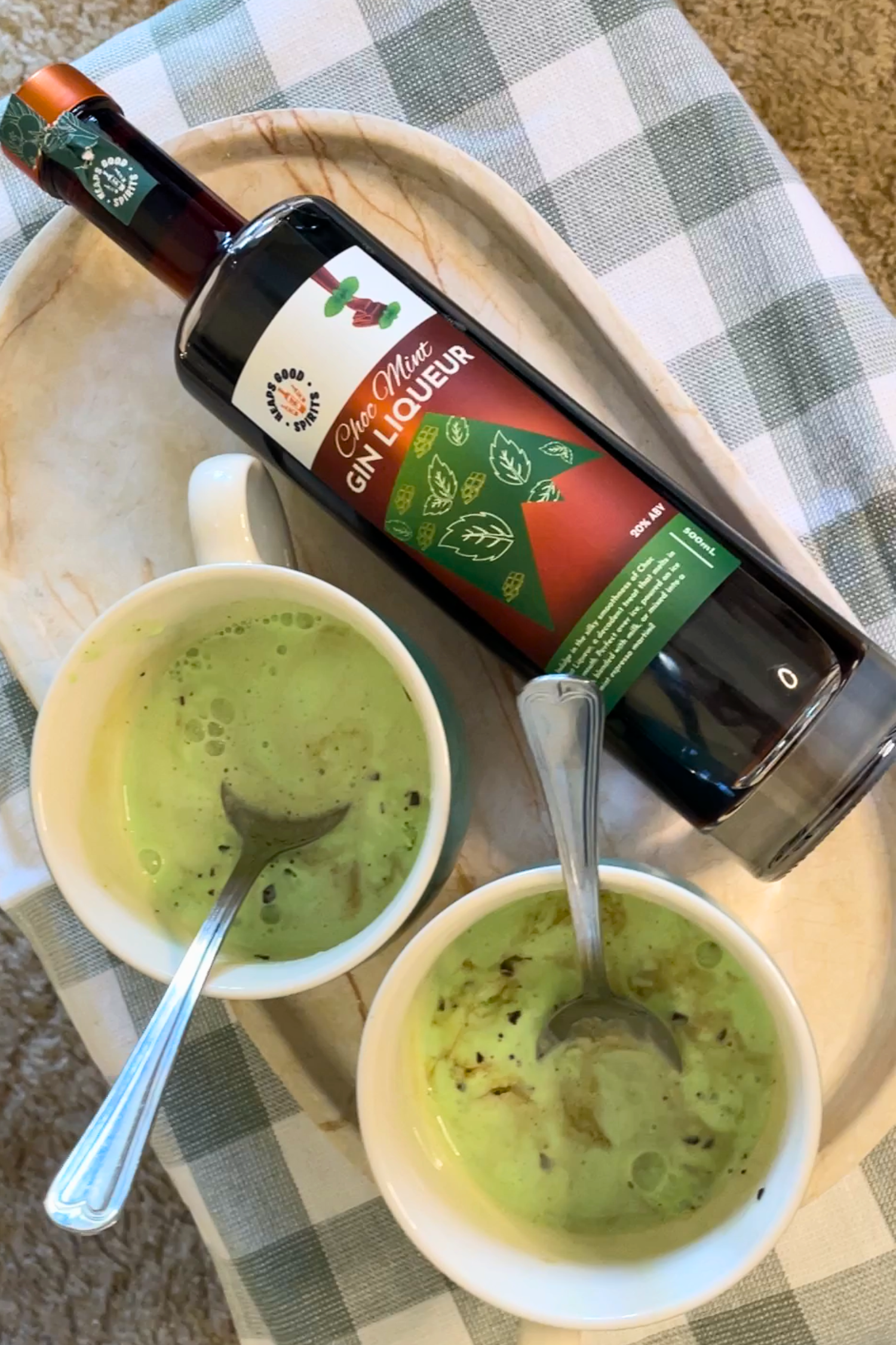 Two bowls of choc mint ice cream with spoons and a bottle of Choc Mint Gin Liqueur on a checkered tablecloth.