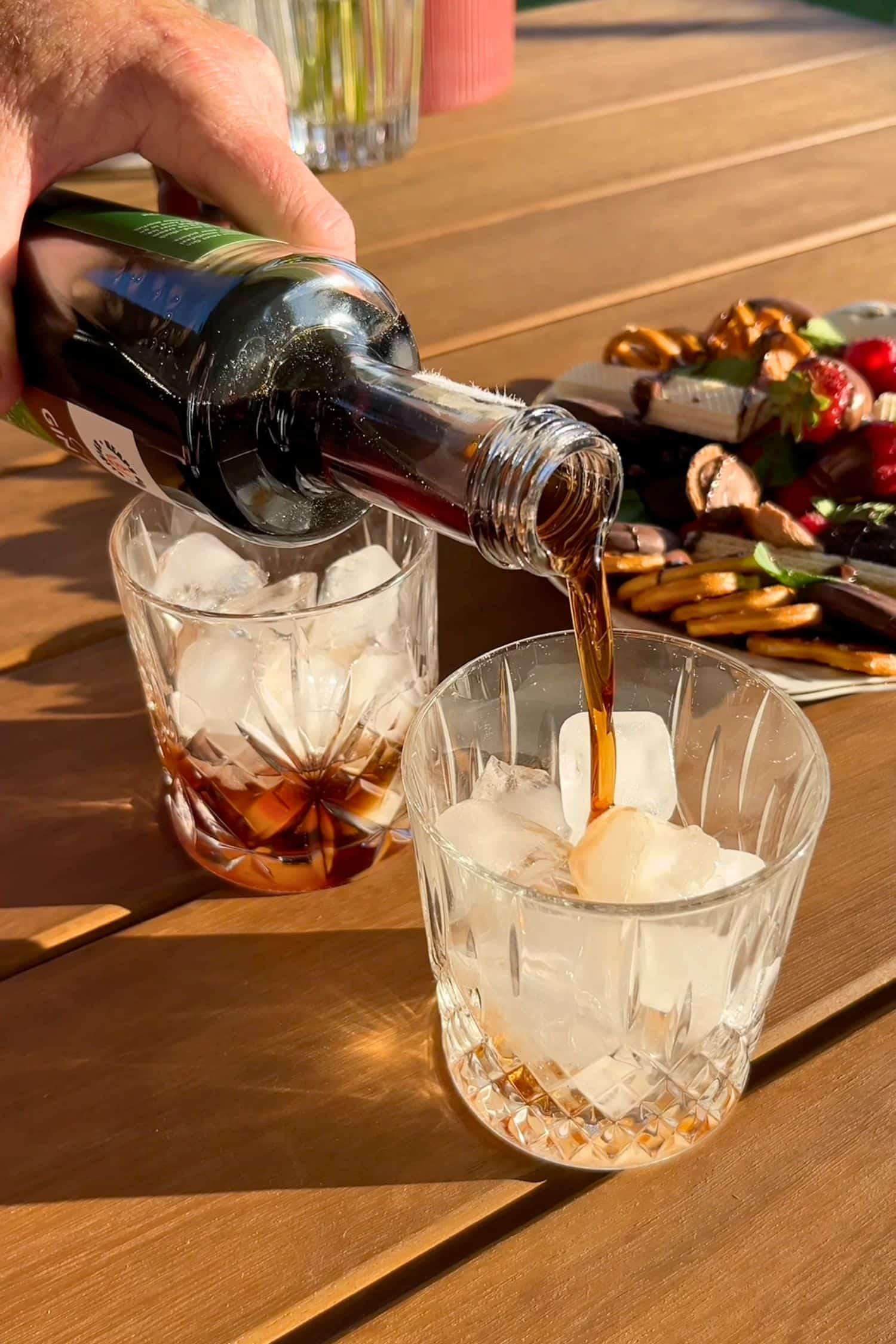Choc Mint Gin Liqueur being poured over ice in crystal glasses outdoors with chocolate and strawberries on grazing board in background