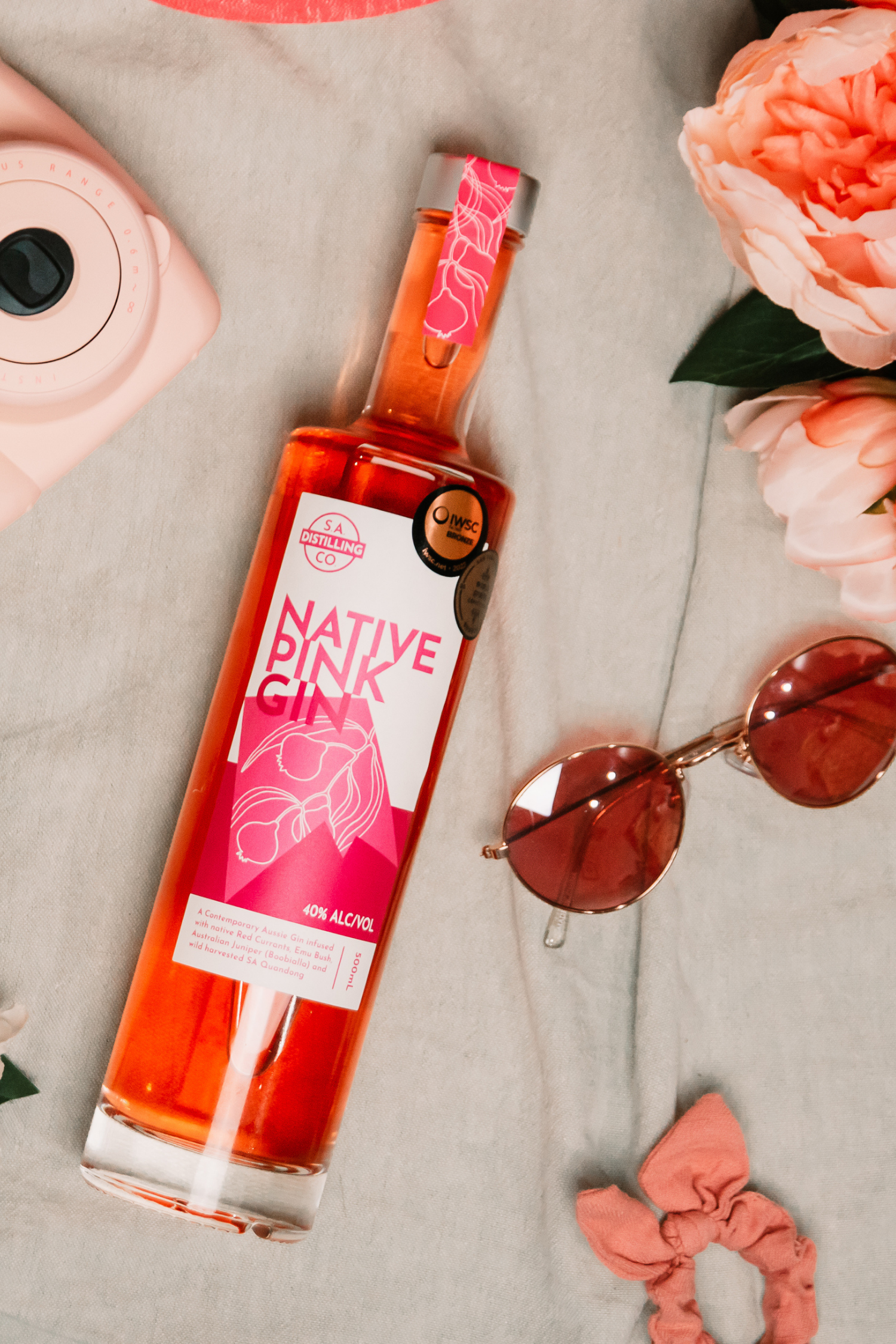 Bottle of Native Pink gin with a pink label, sunglasses, and flowers on a light surface.