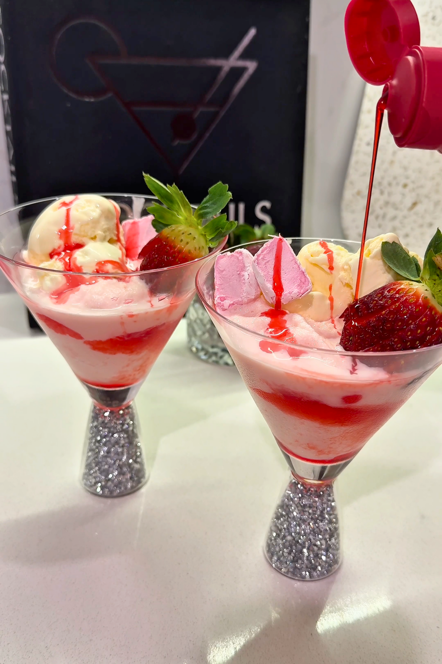 Two dessert glasses with ice cream and strawberries, one being drizzled with red sauce, on a white surface.