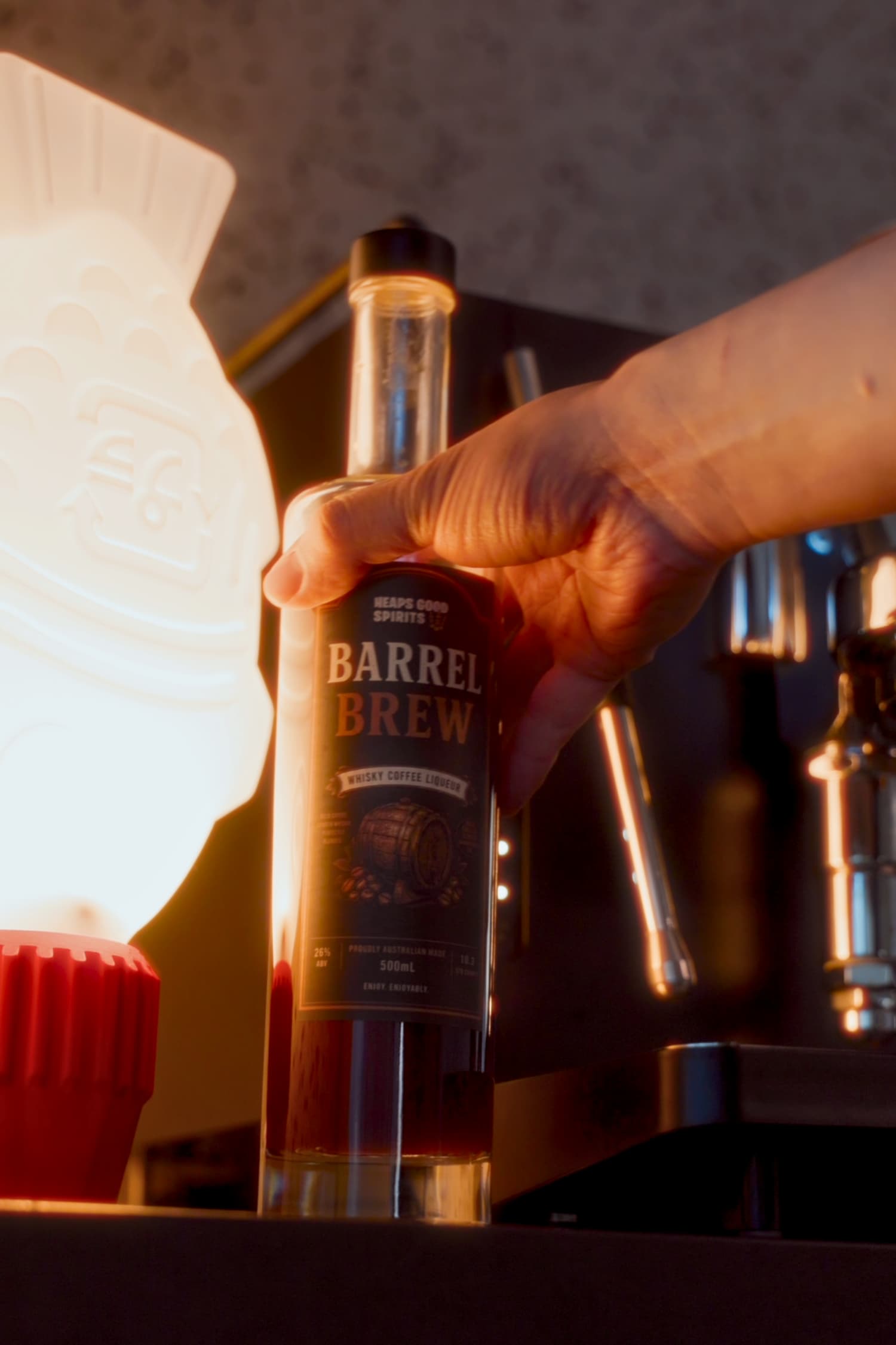 A hand holds a bottle of Barrel Brew whisky coffee liqueur against warm, backlit lighting, with a glowing lamp on the left and an espresso machine blurred in the background.