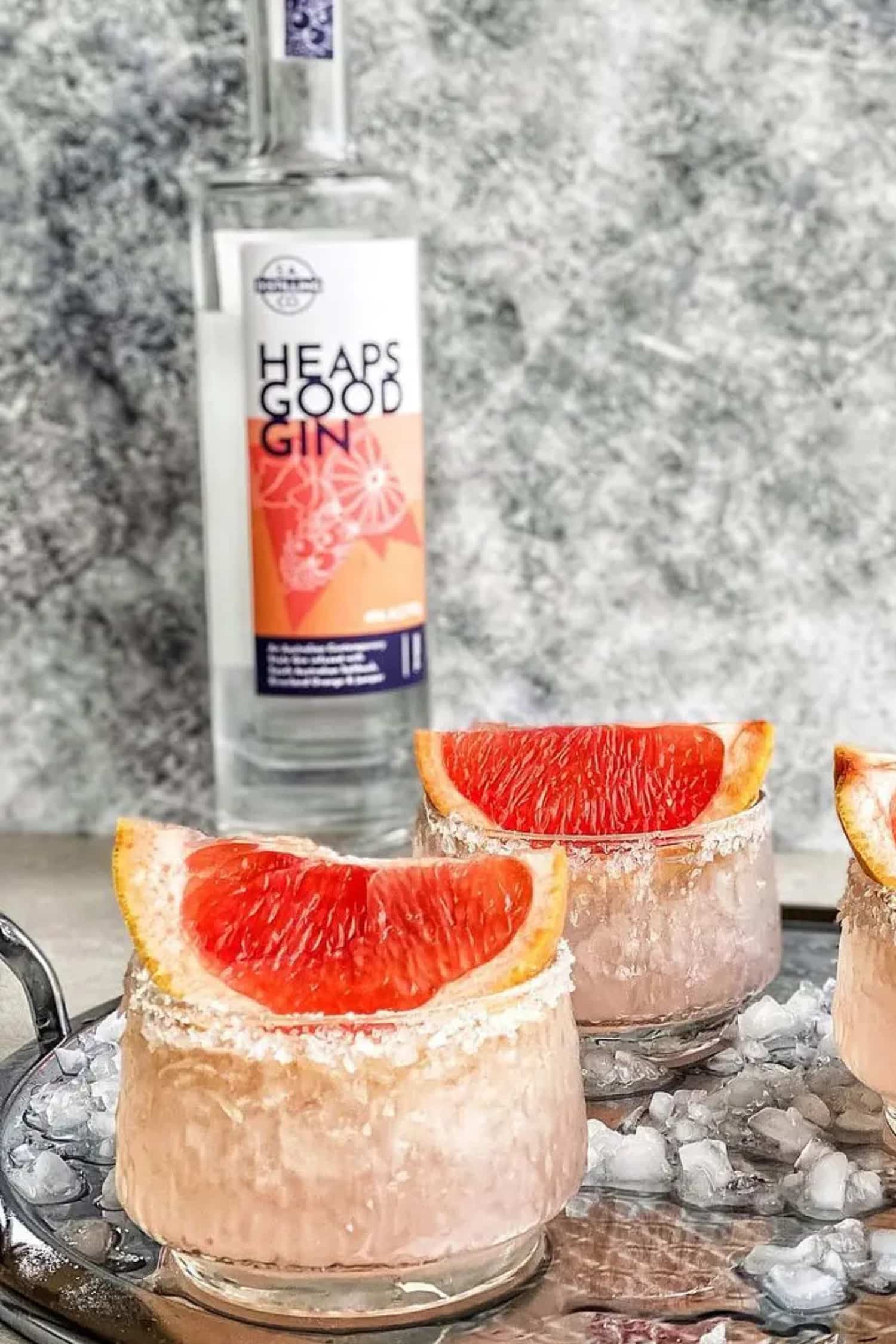 Bottle of Heaps Good Gin with two cocktails garnished with grapefruit slices on a textured surface.