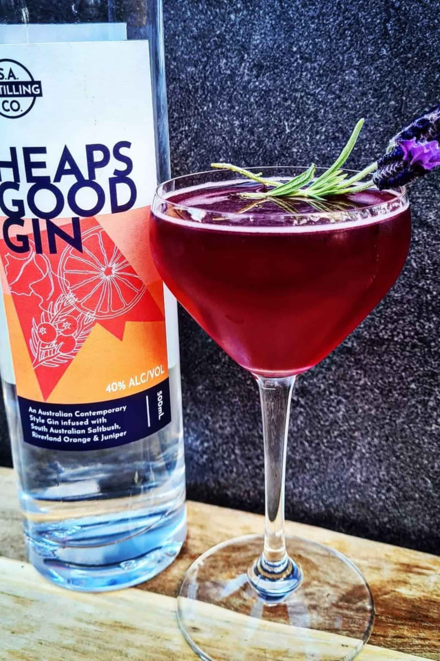 Bottle of Heaps Good Gin next to a cocktail with lavender garnish on a wooden surface.