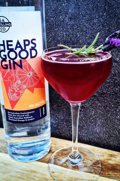 Bottle of Heaps Good Gin next to a cocktail with lavender garnish on a wooden surface.