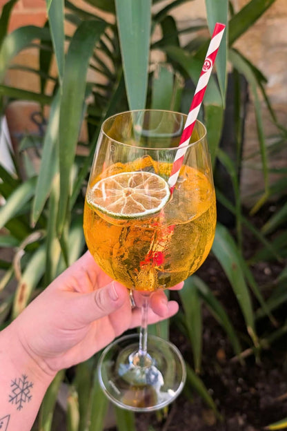 A hand holds a clear stemmed wine glass filled with a Peach &amp; Mango Spritz, garnished with a lime wheel and orange peel, with a red-and-white striped paper straw. Lush green foliage fills the background.