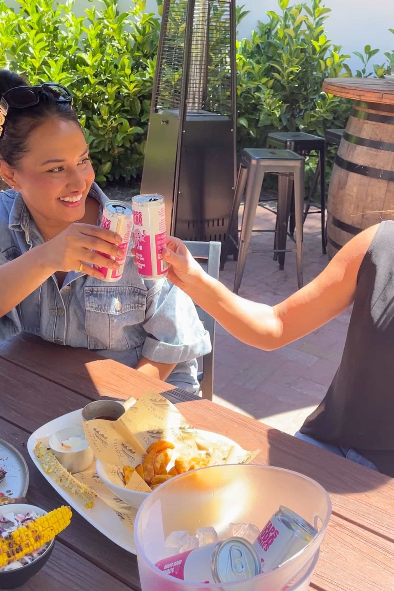 Native pink gin and quandong soda cans being cheersed at outdoor table with food and drinks in sunny setting.