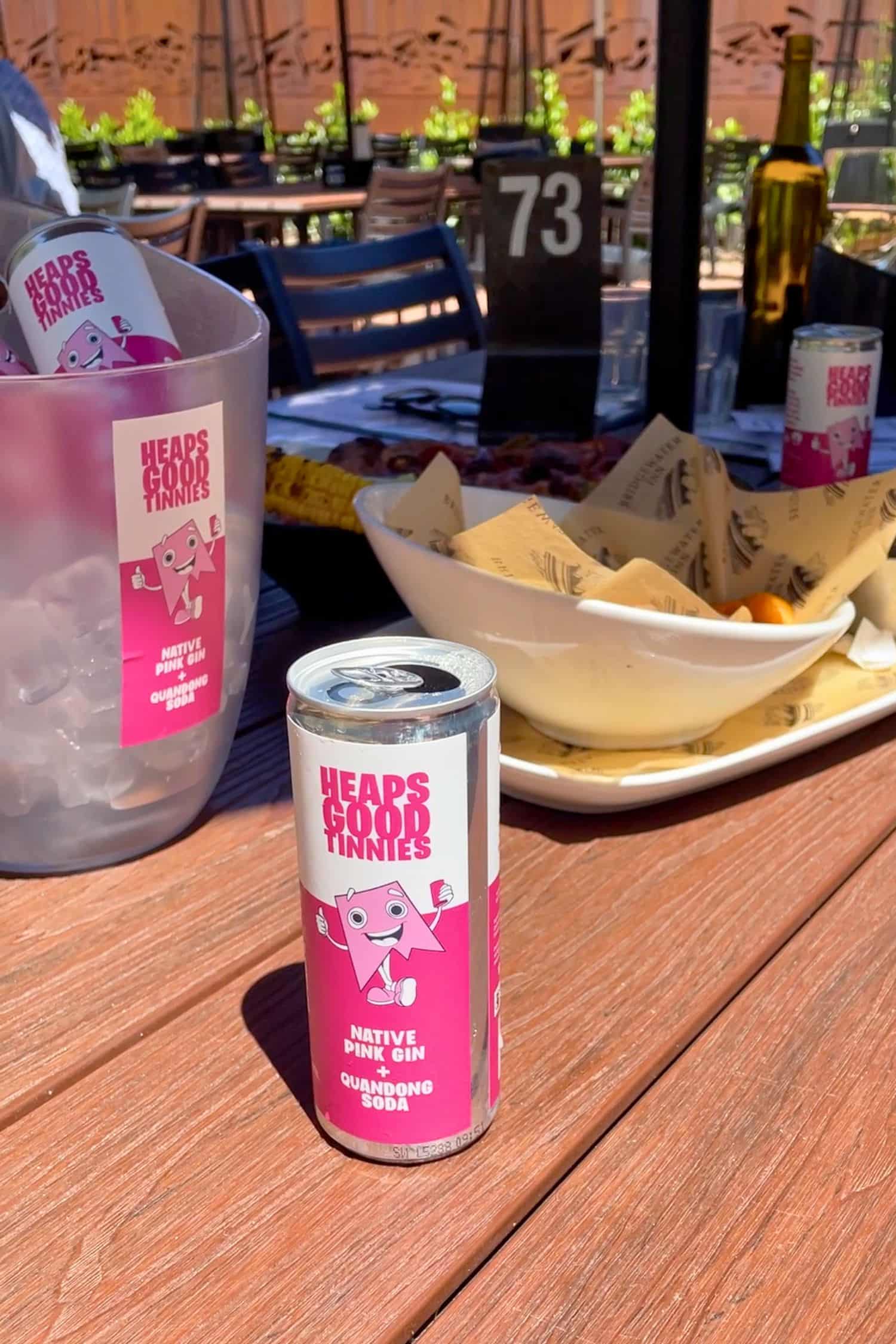 Native pink gin and quandong soda can on outdoor table with ice bucket and food in background.