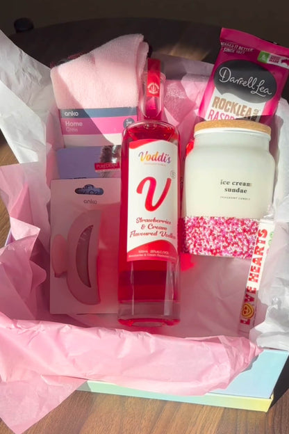 Gift box with a bottle of Strawberries &amp; Cream Vodka, a scented candles, and other items on a wooden surface.