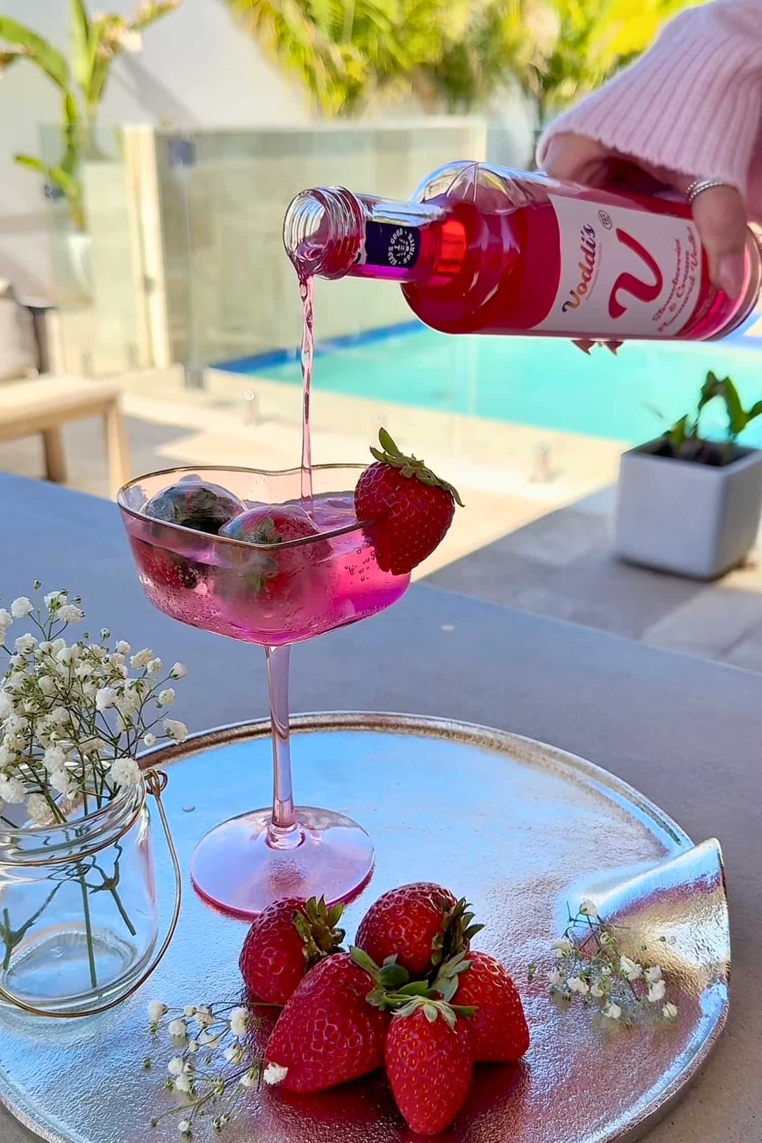 Pink strawberry vodka being poured into a glass over ice with strawberries on a tray, outdoor setting.