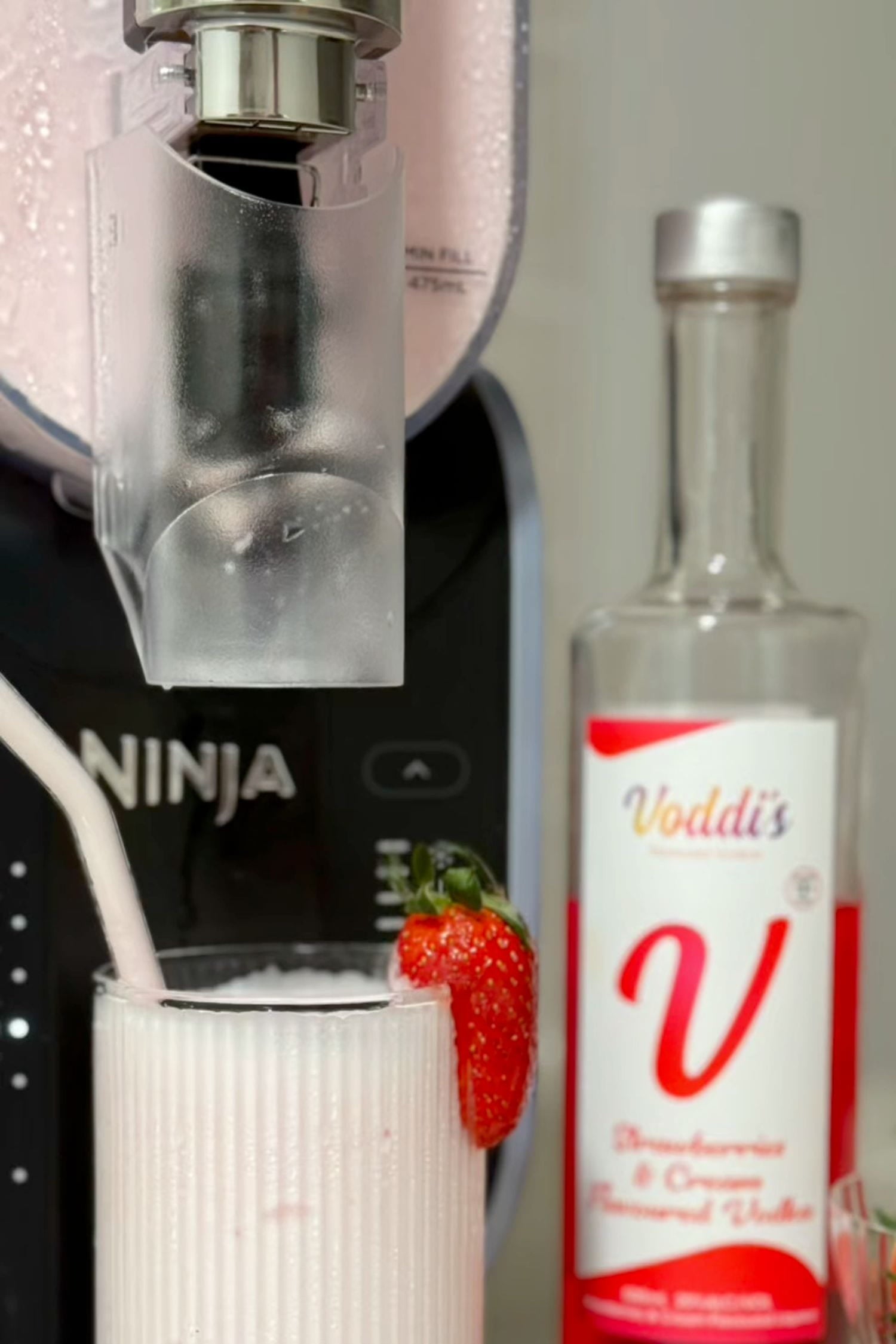 Strawberries &amp; Cream Vodka frozen slushie cocktail with fresh strawberry garnish beside Voddi’s bottle and Ninja slushie machine.