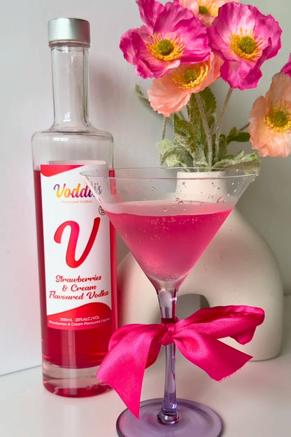 Strawberries &amp; Cream Vodka pink martini cocktail in glass with ribbon bow, Voddi’s bottle and pink flowers.