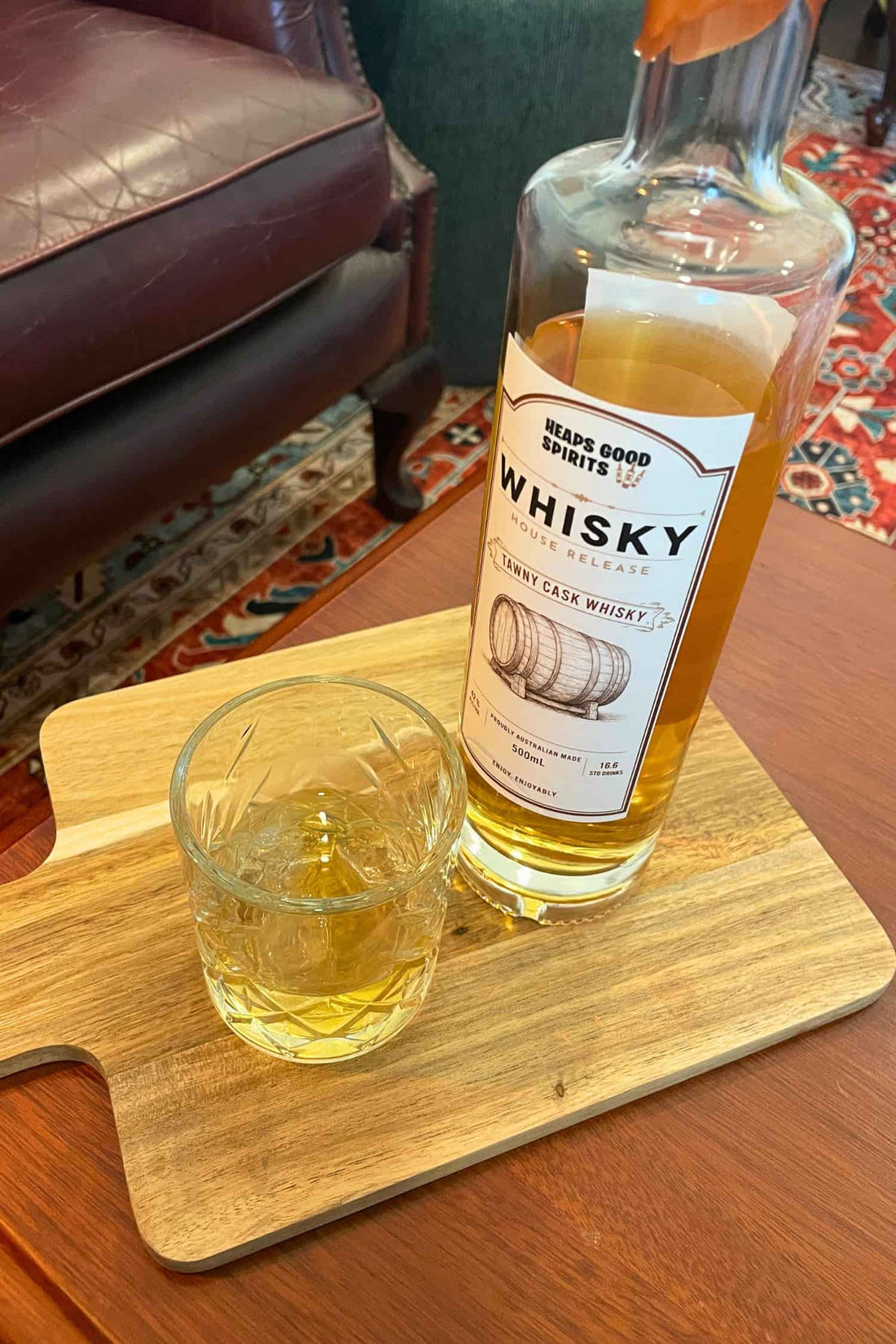 Bottle of Tawny Cask Whisky on a table, with a glass of whisky on the rocks. In the background is a cosy lounge setting.